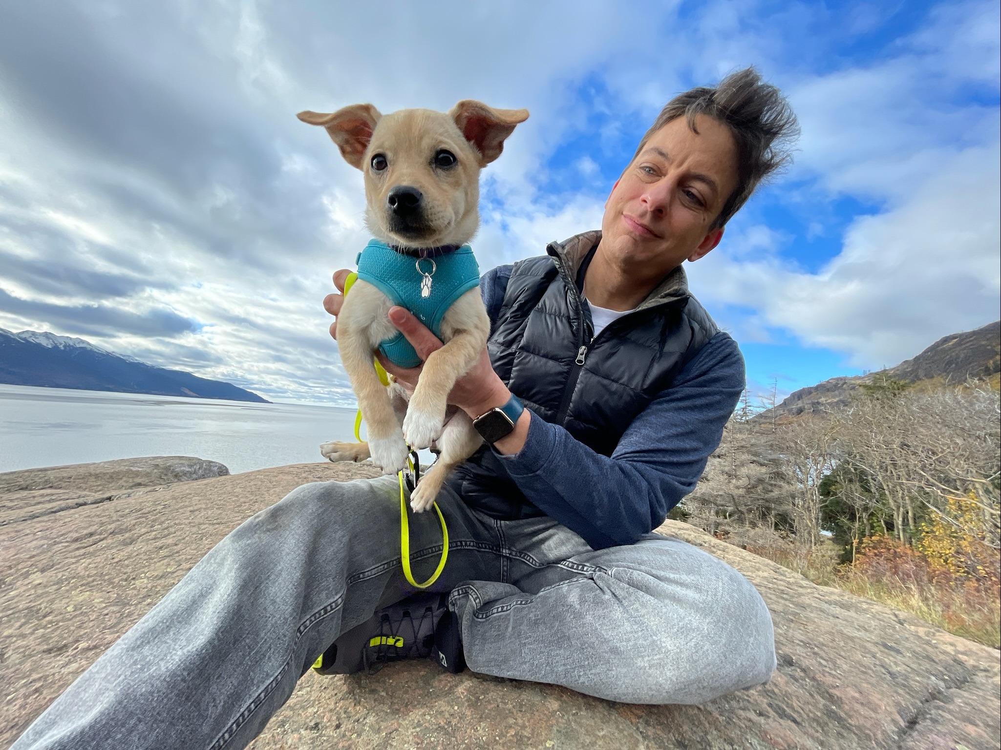 Zak holding Biscuits, a small yellow Chihuahua mix in a blue harness, beside water and mountains in Alaska.
