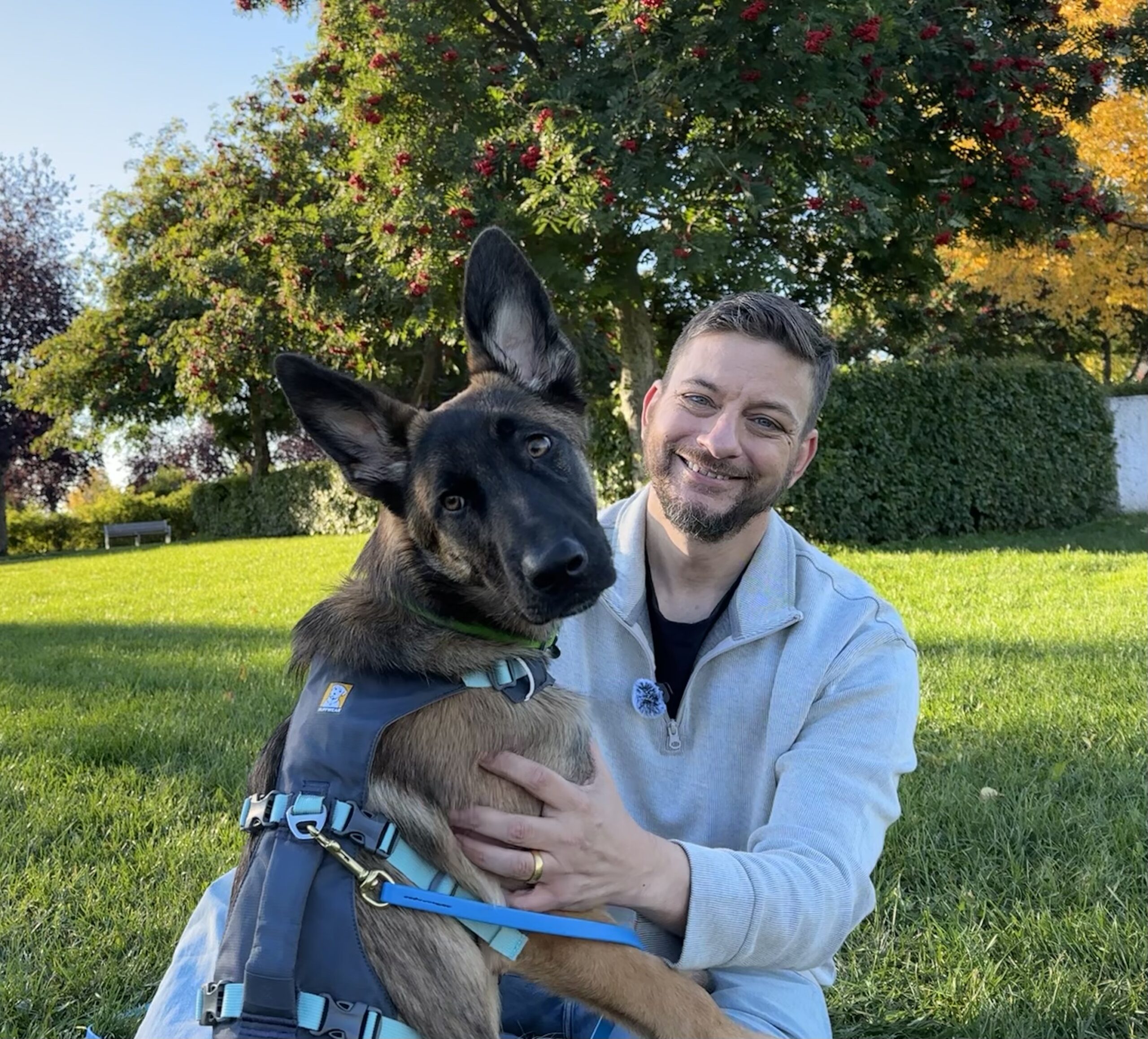 Zak sitting with Wallace, a Malinois mix with large upright ears, beneath autumn trees.